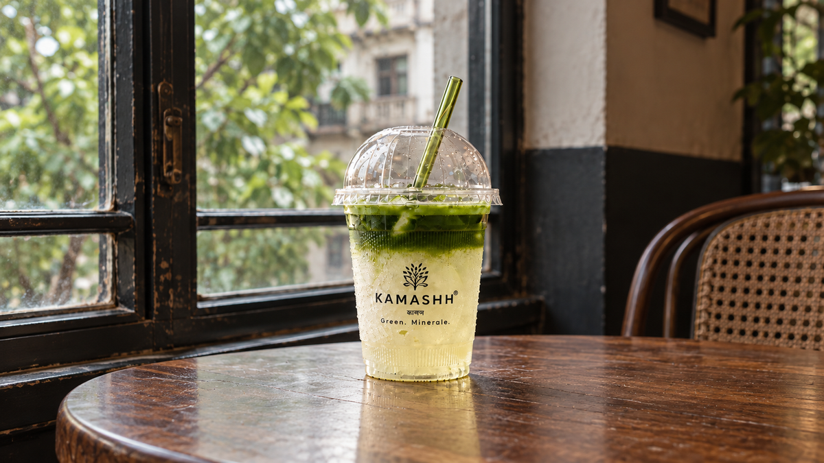 Kamashh iced matcha latte in a branded takeaway cup with a green straw, on a wooden café table beside a window.